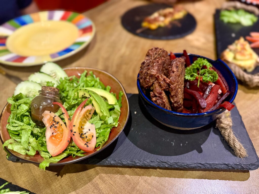 A restaurant dish at El Sol Monteverde featuring a fresh green salad with sliced tomatoes, avocado, cucumber, and black sesame seeds alongside a vegetarian entre.